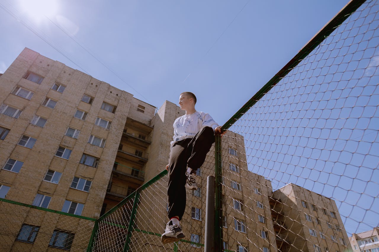 Teenager sitting atop a chain-link fence with urban buildings in the background under a clear sky.